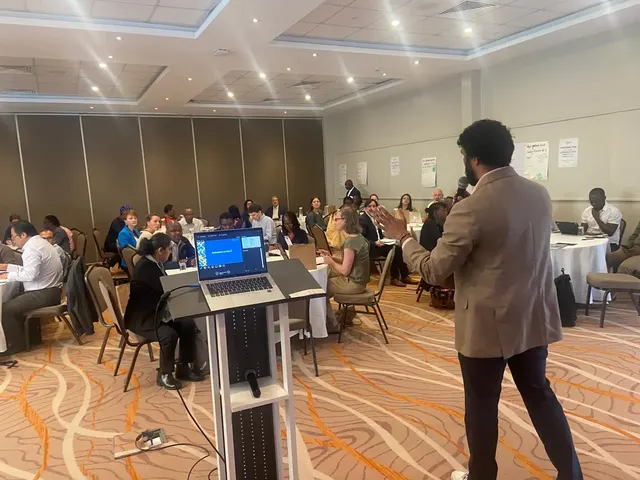 Speaker holding a microphone during a workshop with participants seated around tables.